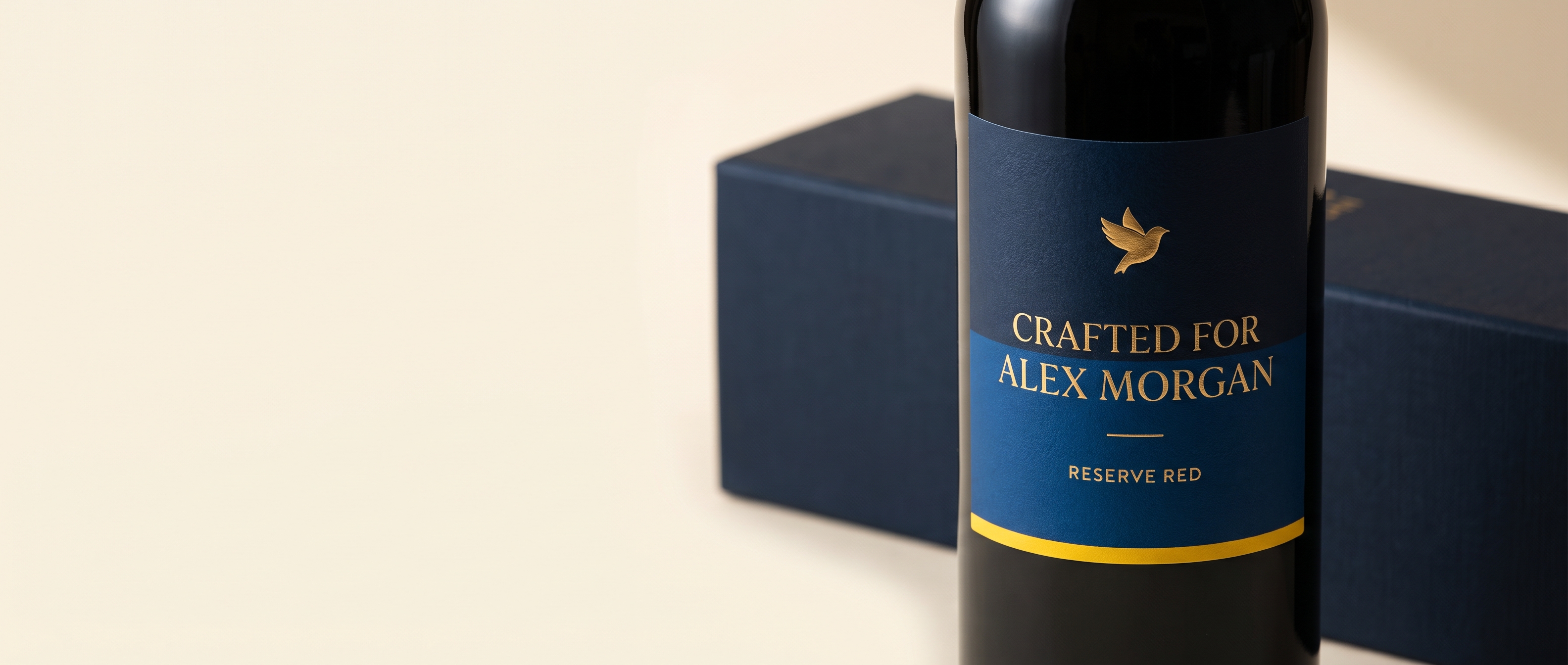 Close-up of a personalised navy wine label reading 'Crafted for Alex Morgan' in gold, beside its navy gift box on a warm cream backdrop